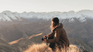 Man atop a snow capped mountain holding a camera wearing a beanie by Will & Bear