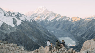 Three friends sit on a rocky peak, overlooking a snowy mountain range, each wearing a snow hat by Will & Bear.